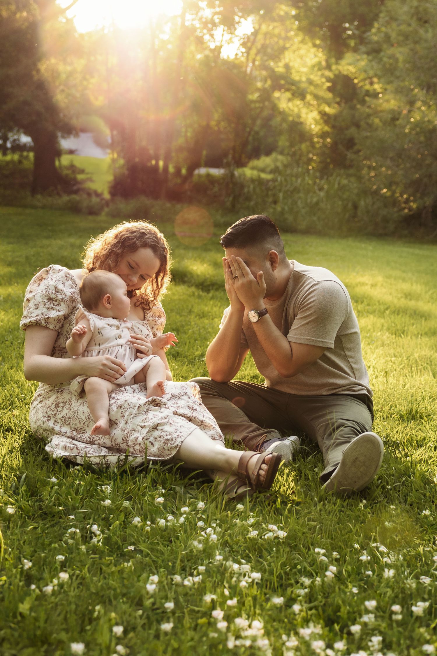 Family sitting on grass in sunny park, mother holding baby in floral dress while father covers face with hands.