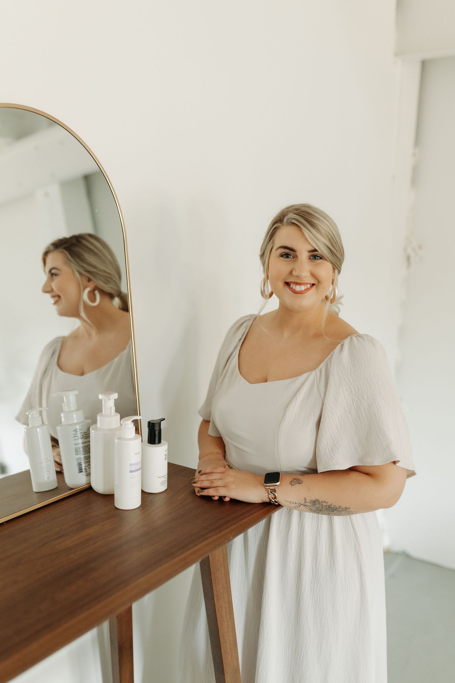 Smiling blonde woman in white dress stands beside wooden vanity with skincare products and arched mirror.