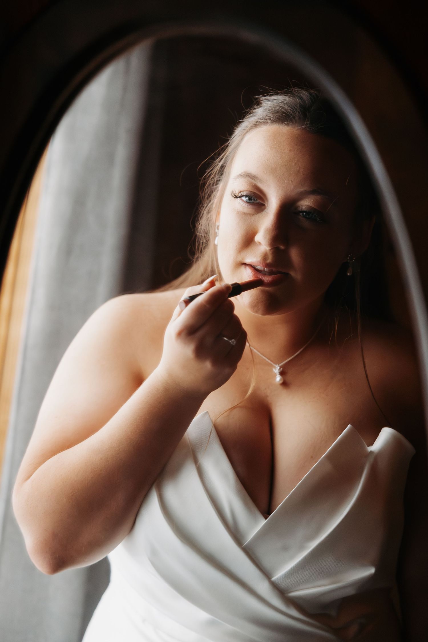 Bride in white strapless gown applies lipstick while gazing into an oval mirror on her wedding day.