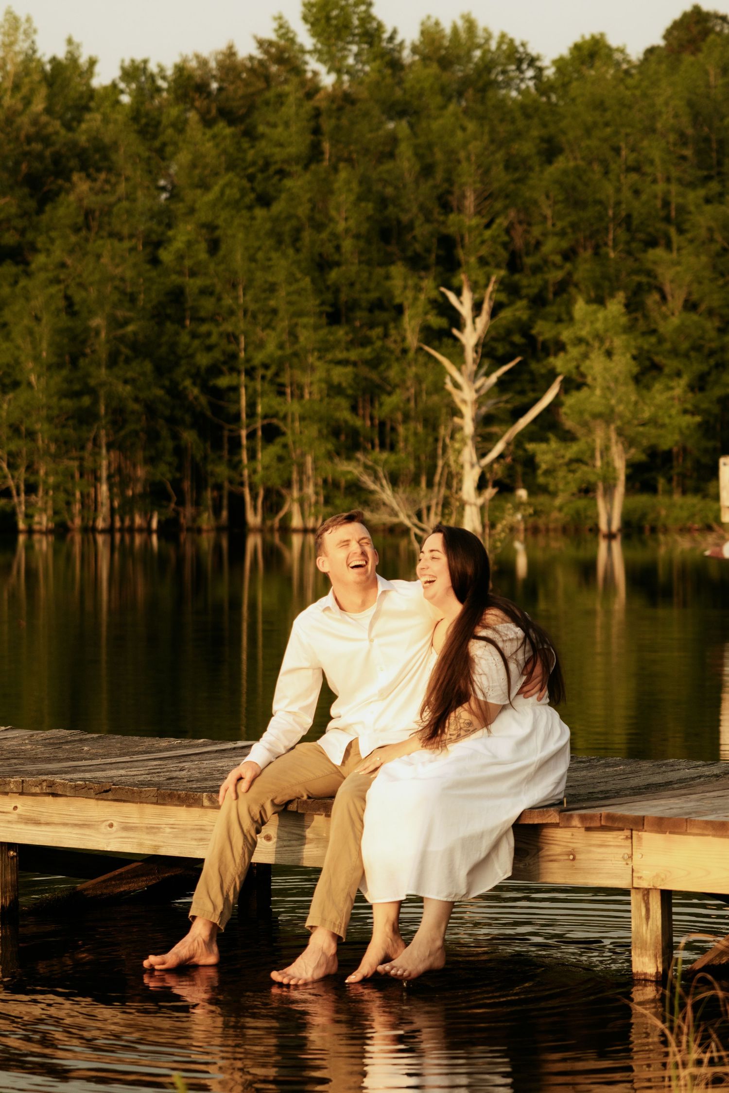 A laughing couple sits barefoot on a wooden dock by a calm lake surrounded by lush green trees at golden hour.
