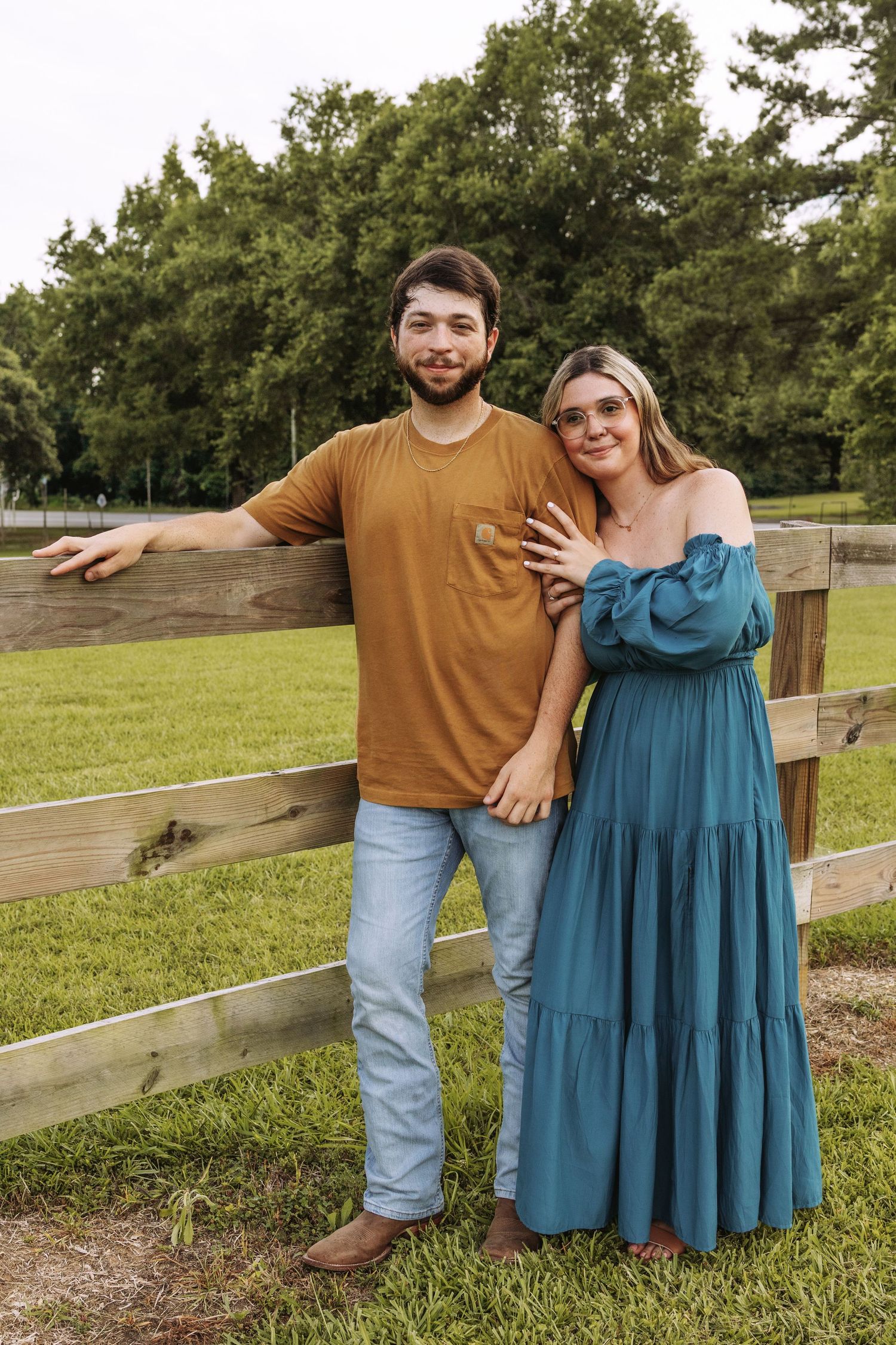 A couple stands together by a wooden fence in a grassy outdoor setting wearing casual clothing.