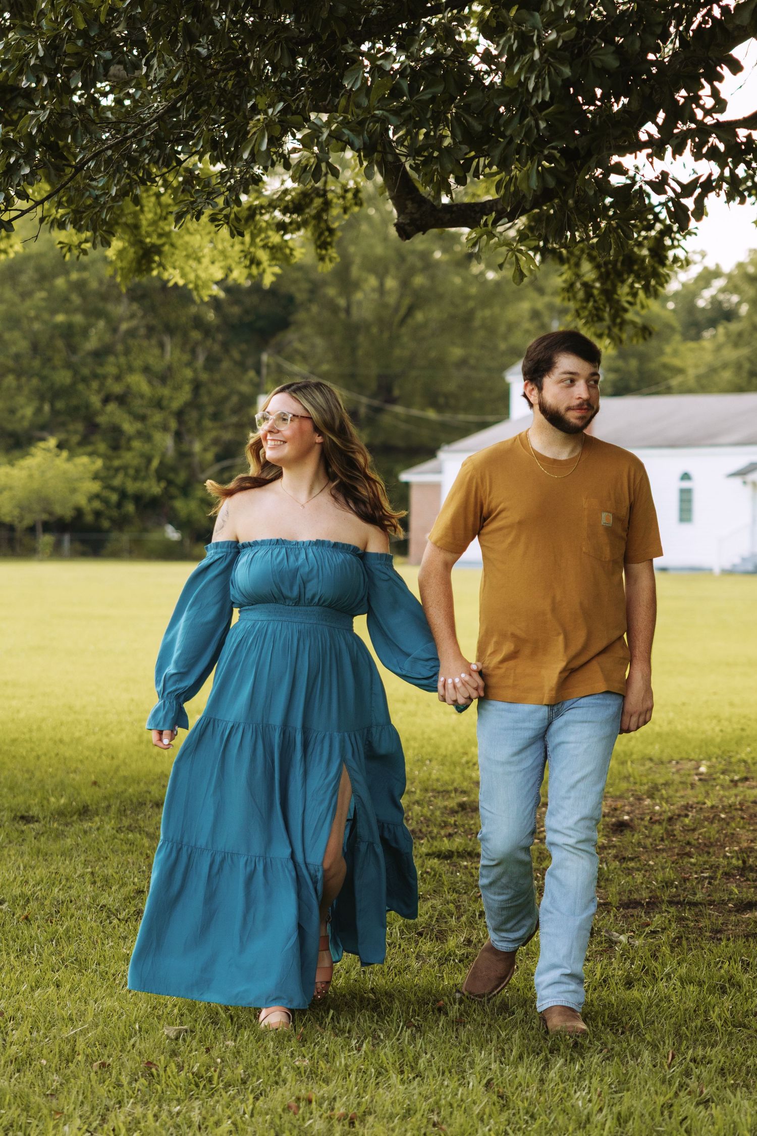 A couple walks hand in hand through a grassy field on a sunny day, her in a blue dress and him in a yellow shirt.