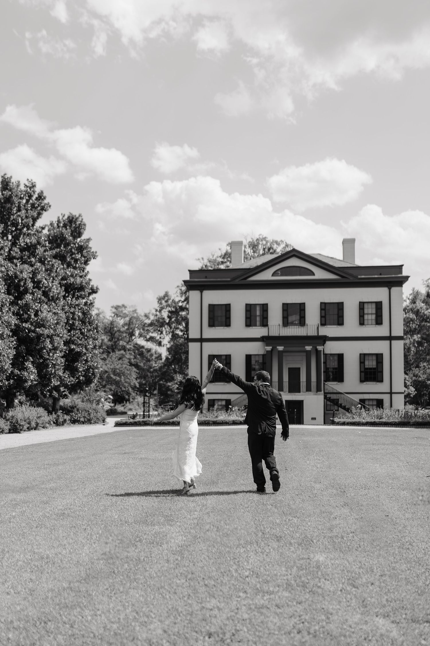 Black and white photo of a couple holding hands walking toward a grand historic mansion on a lush green lawn.