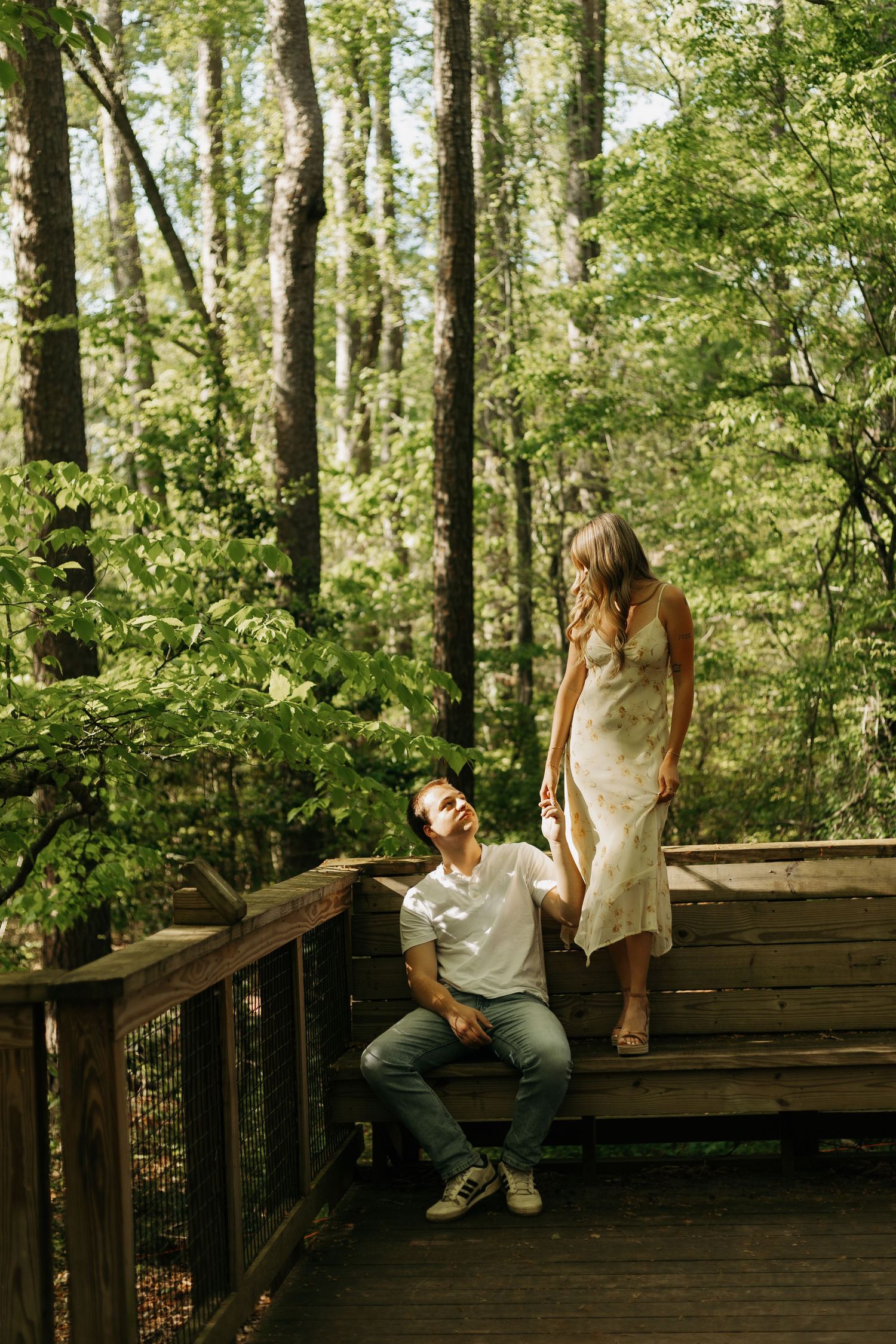 Couple on a wooden forest boardwalk, man sitting against railing, woman in floral dress standing nearby.