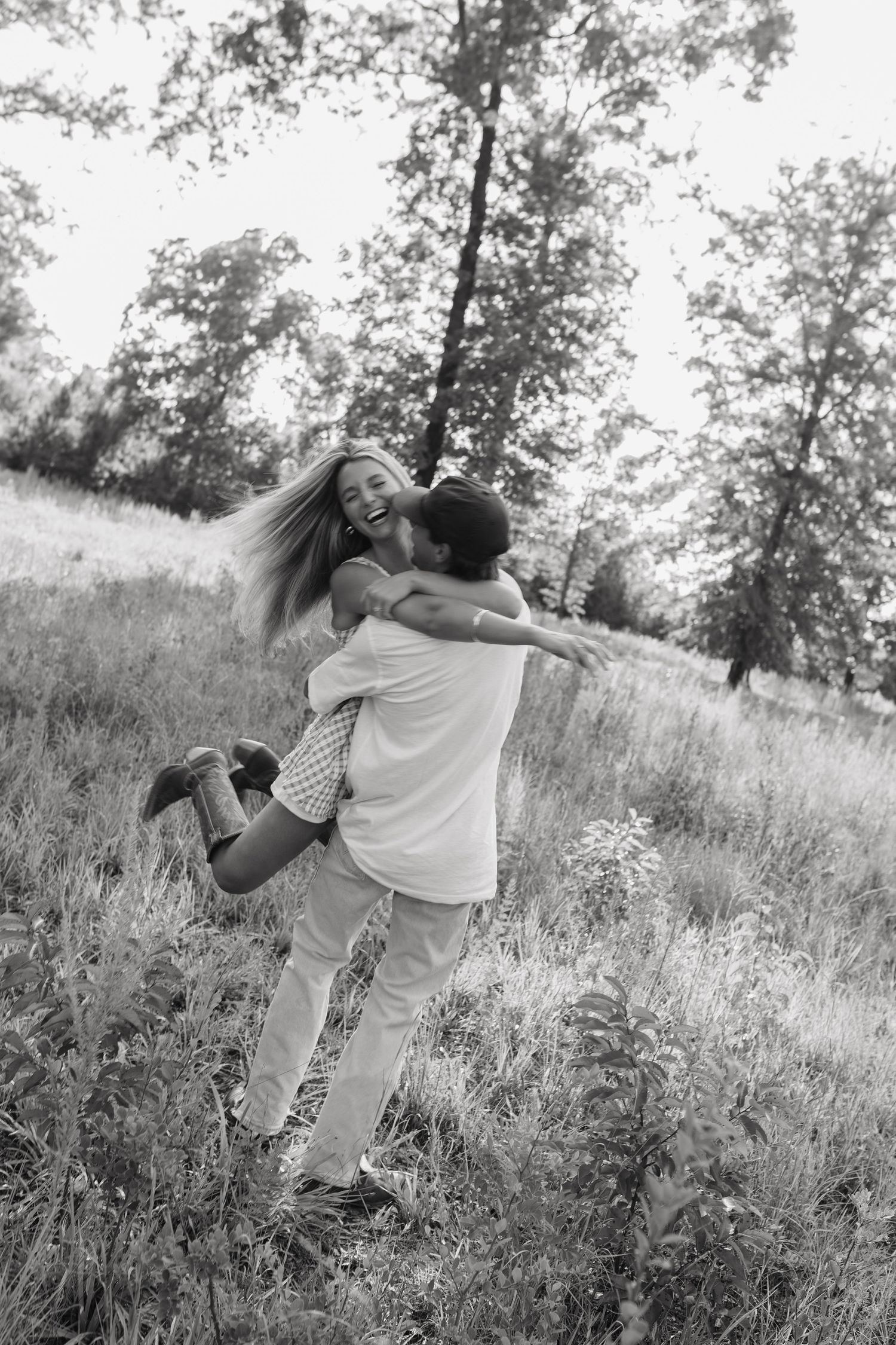 Black and white photo of a joyful couple embracing in a grassy field, man lifting woman as she laughs with trees behind.