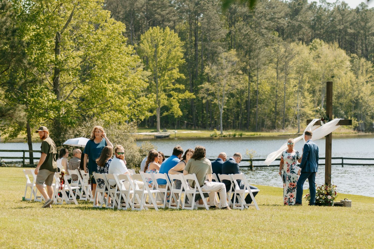 Outdoor wedding ceremony on green lawn near lake with white chairs and guests gathering among trees on sunny day.