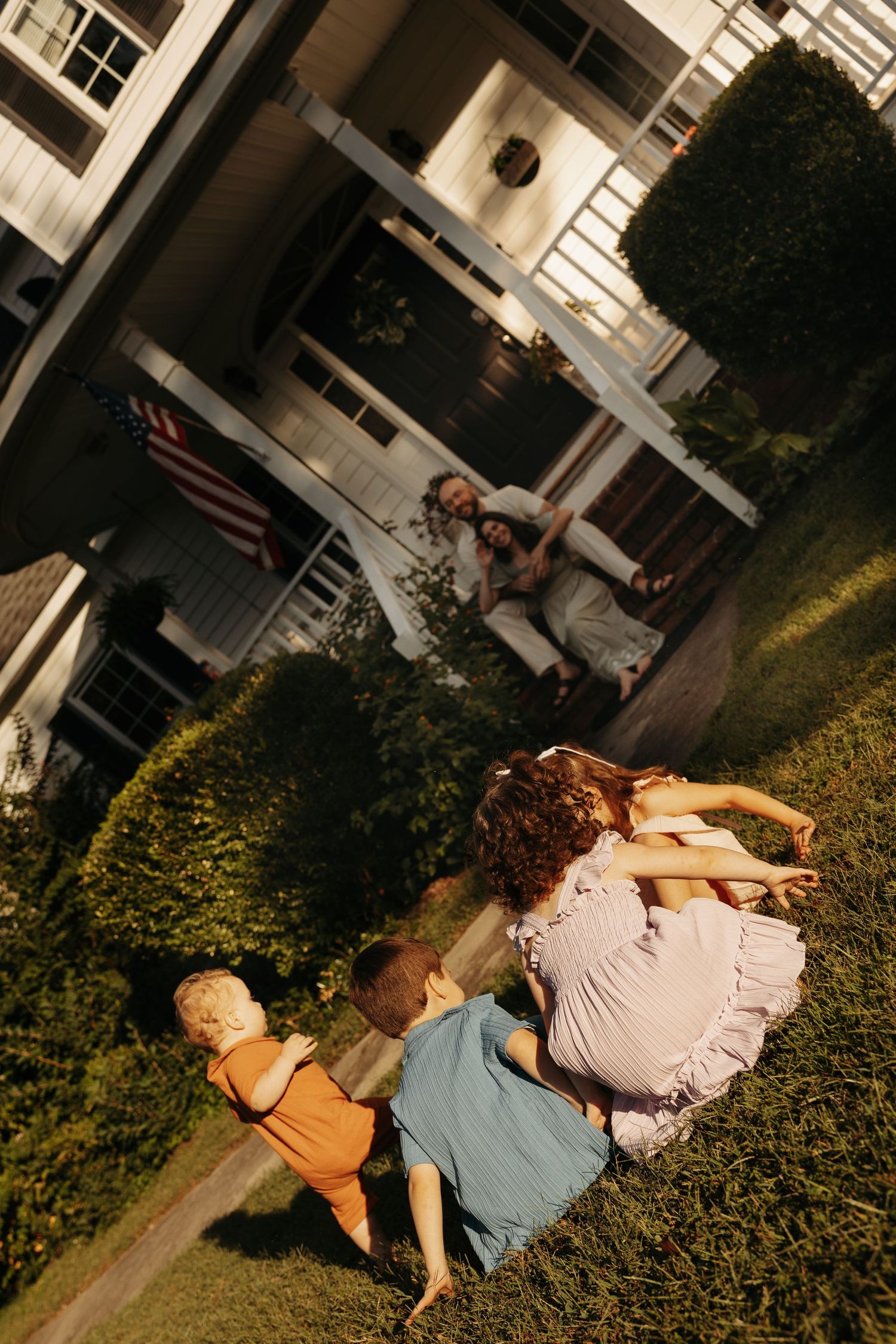 Children sitting on grass in front of a white house with an American flag, watched by an adult on the porch.