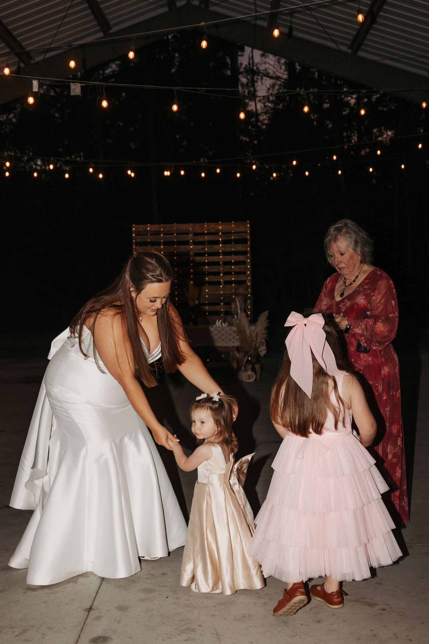 Bride in white gown bends to hold hands with flower girls in pink dresses at outdoor evening wedding reception.