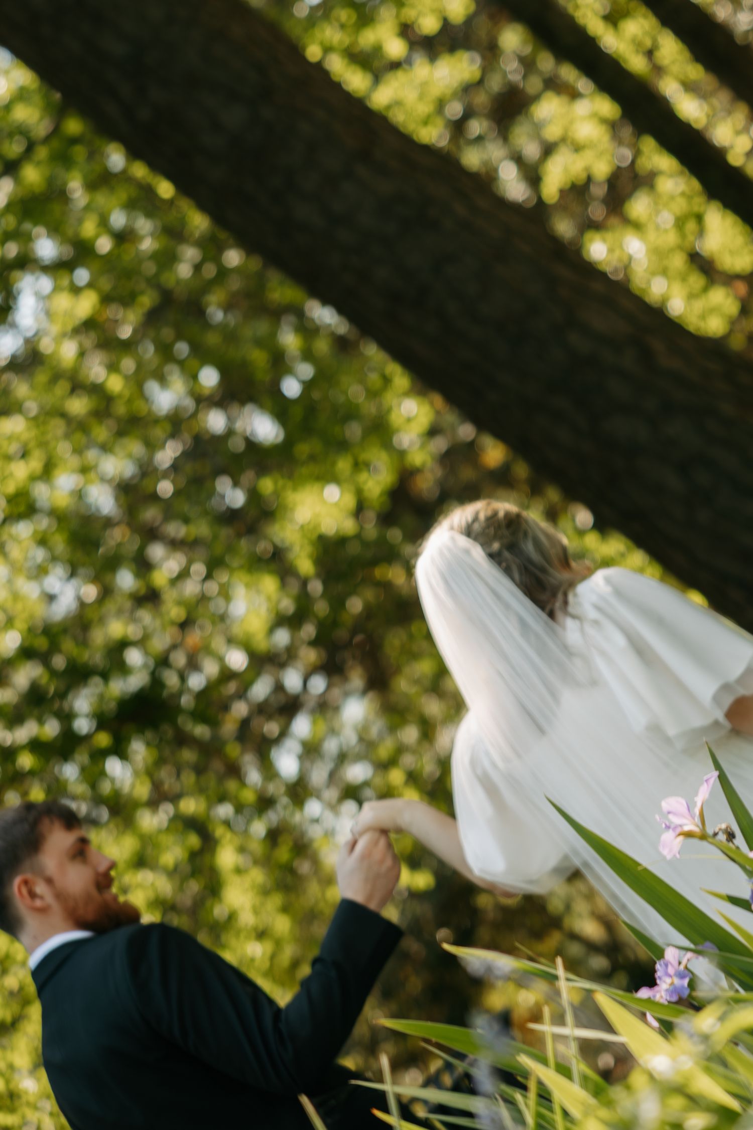 Groom holds bride's hand as she walks ahead in a lush green garden, her white veil and dress flowing gracefully.