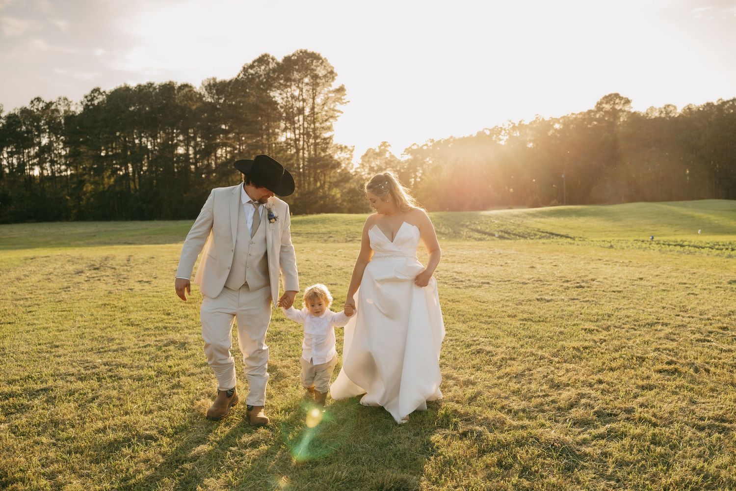 Bride and groom in wedding attire walk with toddler child across sunlit green field at golden hour.