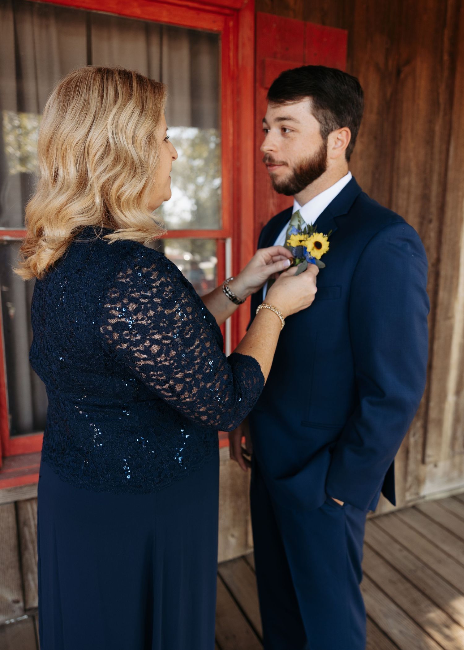 Woman in navy lace dress pinning a sunflower boutonniere on a man in a navy suit before a wedding.