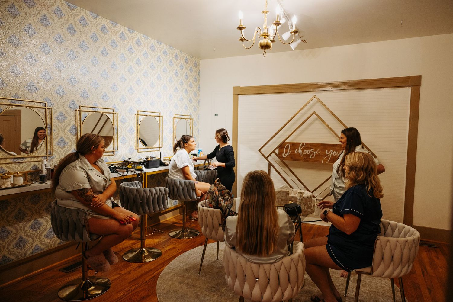 Women gathered in a elegant beauty salon with gold mirrors, chandelier, and decorative wallpaper for a group appointment.