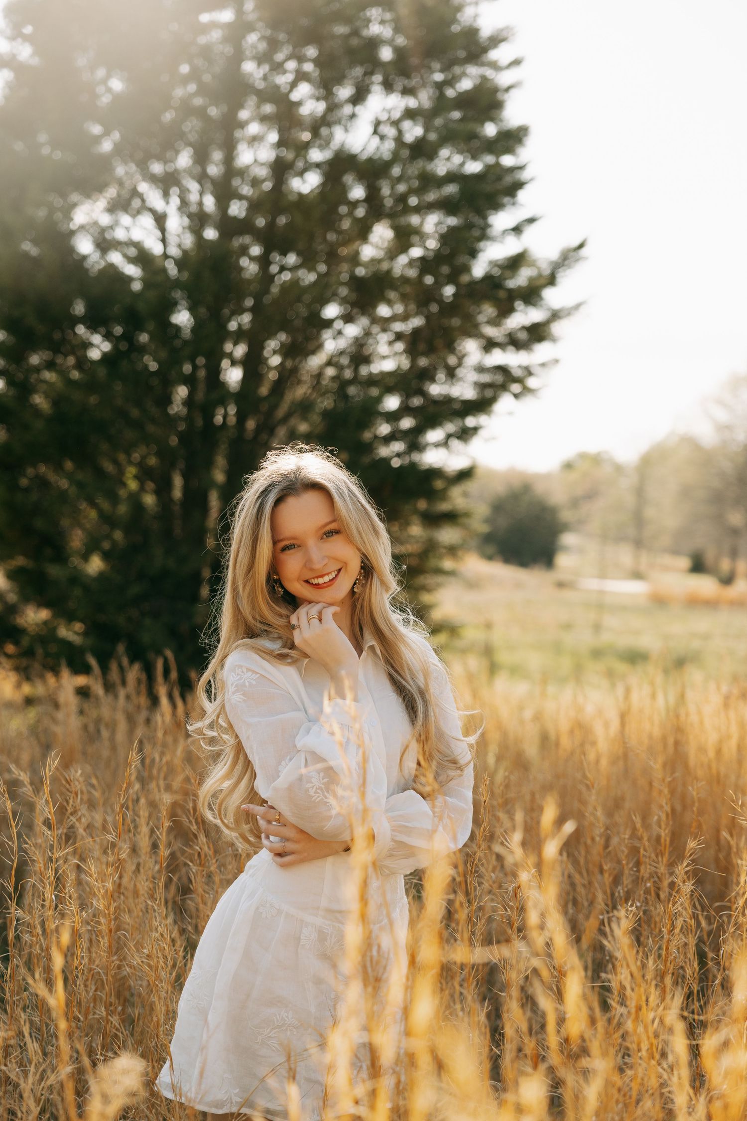 Young blonde woman in white dress smiling in golden field with trees in background on sunny day.