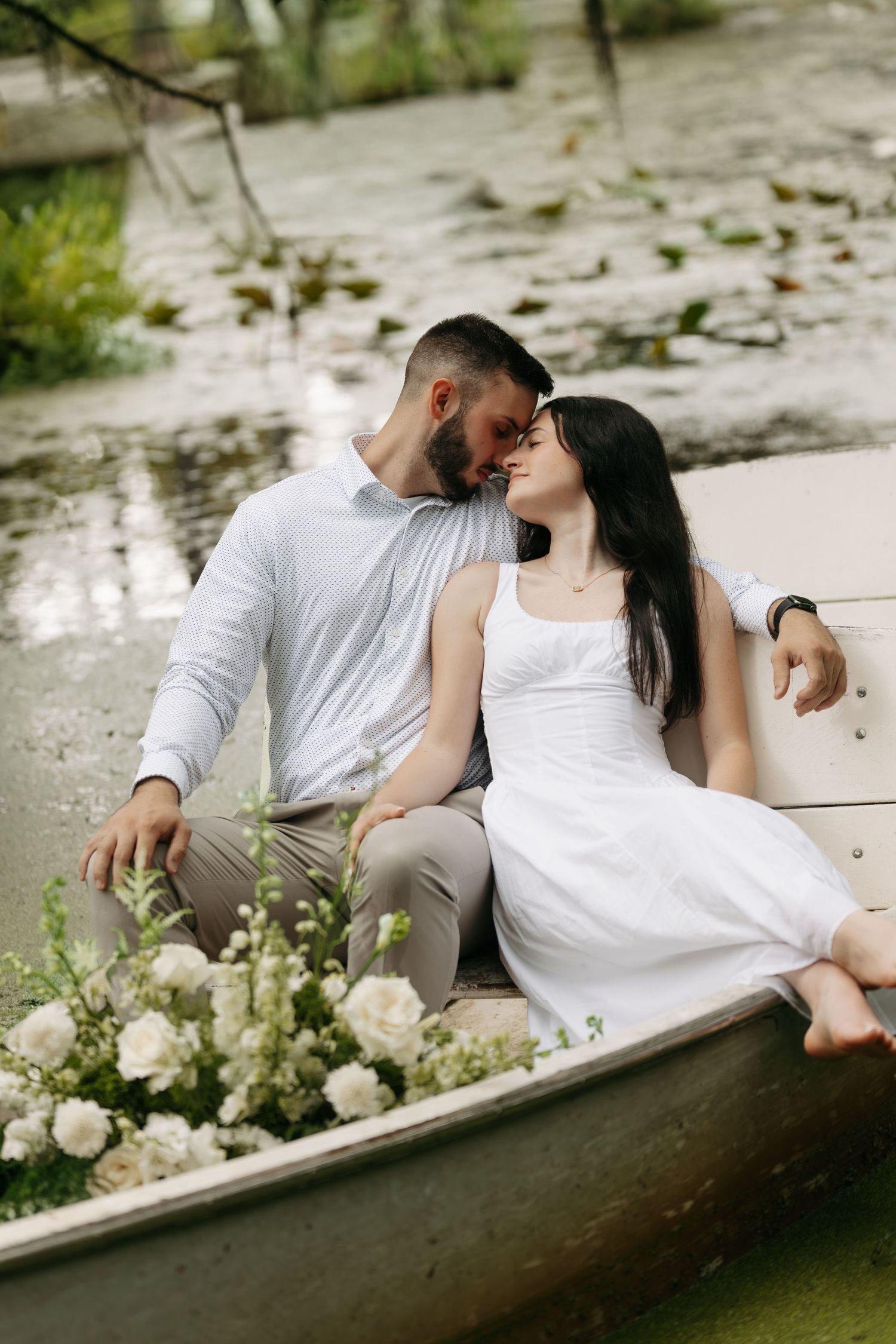 Romantic couple in white outfits sharing an intimate moment in a flower-adorned boat on a serene pond.