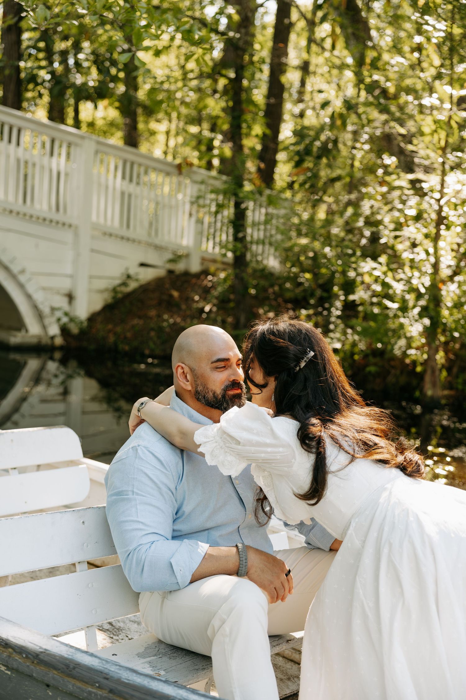A couple shares a romantic moment on a white bench near a bridge surrounded by lush greenery.