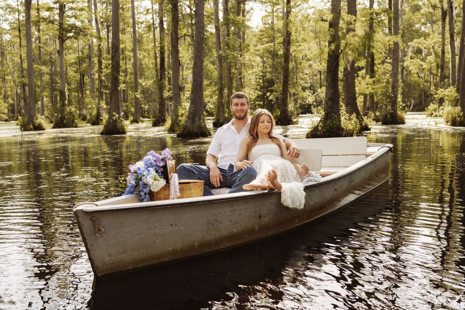 Couple sitting in a canoe on a serene cypress swamp, surrounded by lush green trees and purple flowers.