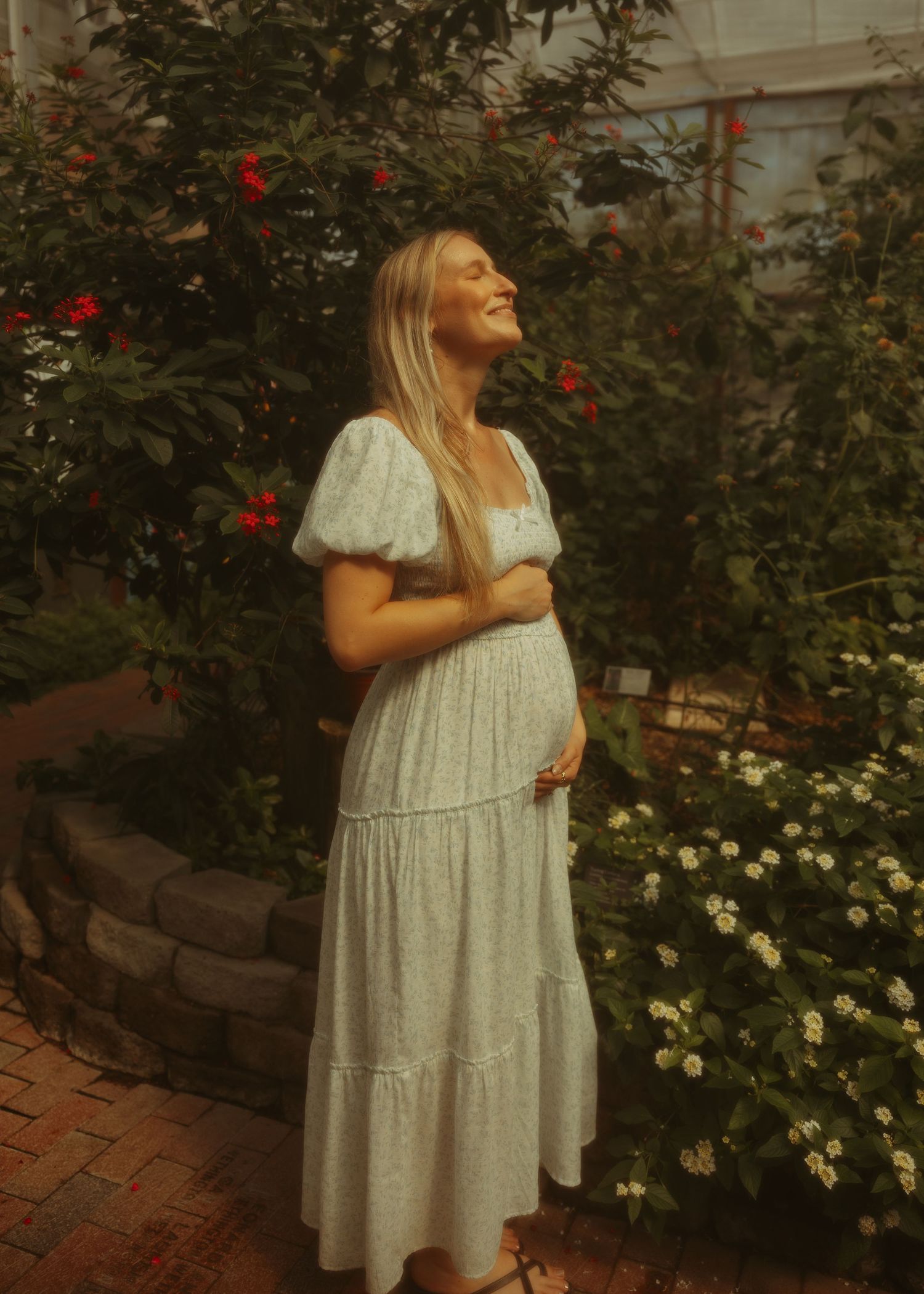 Smiling pregnant woman in white floral maxi dress standing among blooming red and white flowers.