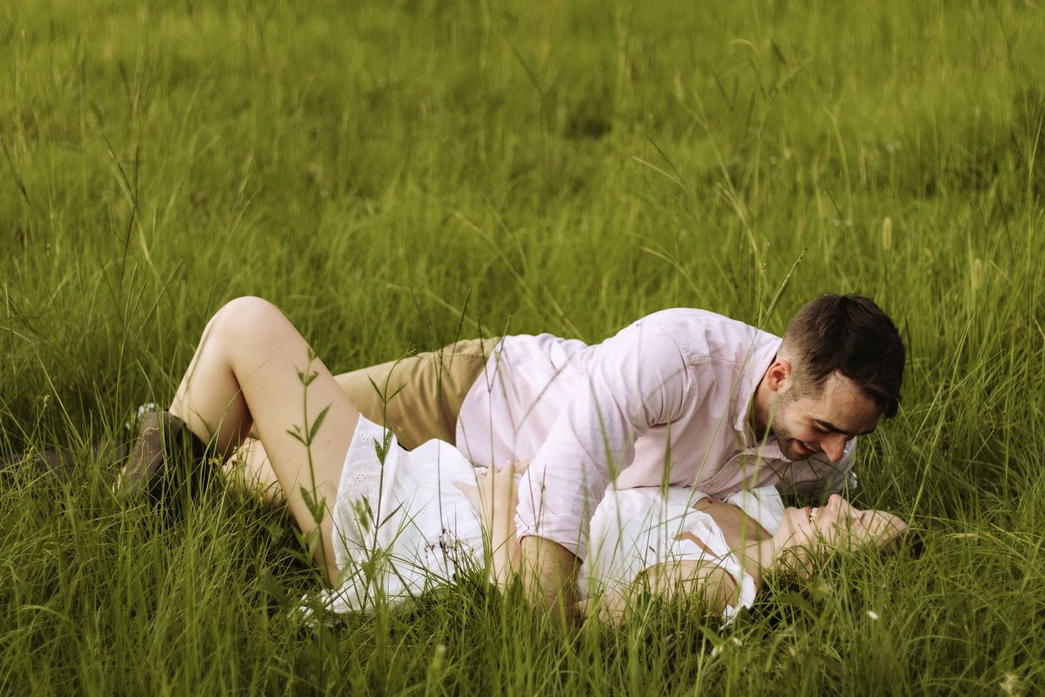 A couple in casual summer attire lies together on tall green grass in a meadow.