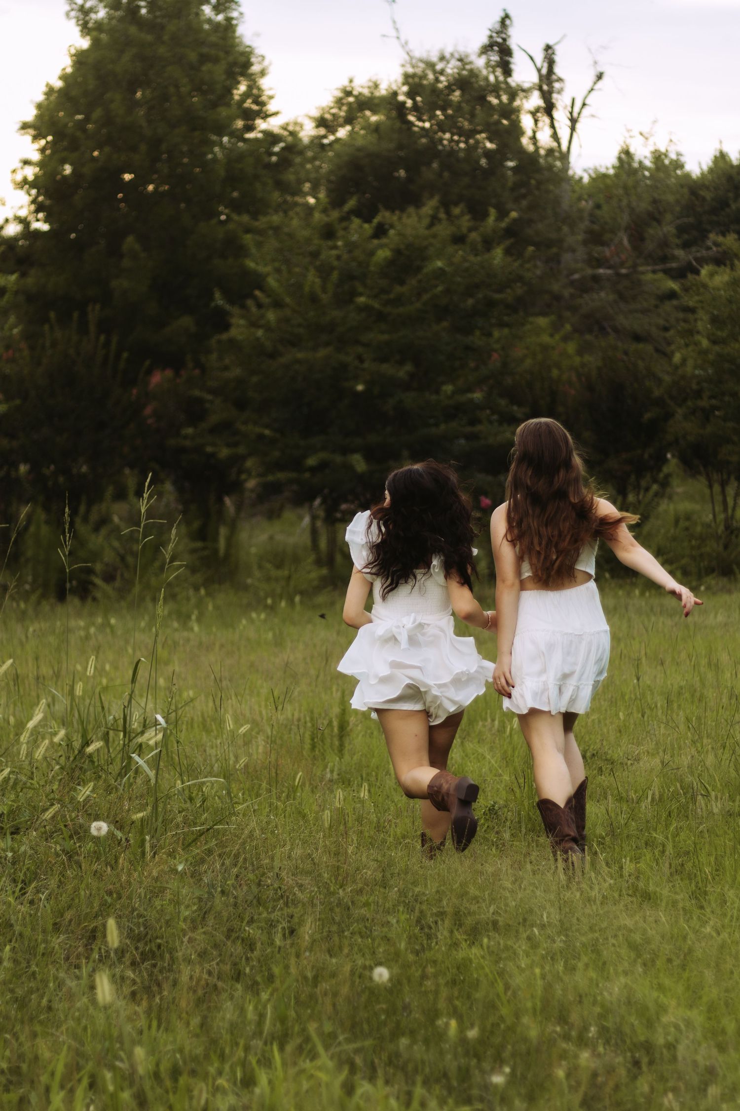 Two people in white dresses and boots run through a green summer meadow with trees in the background.