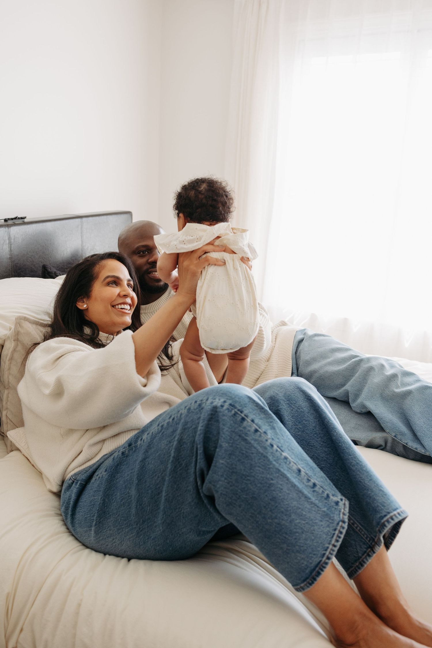 Happy interracial family with mother, father, and baby relaxing together on a white bed in a bright bedroom.