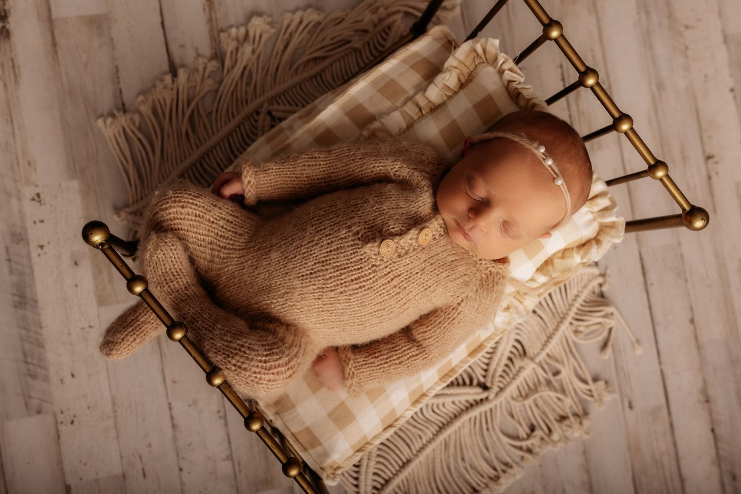 Newborn baby sleeping peacefully in a tiny bed wearing a knit tan romper with a delicate headband.