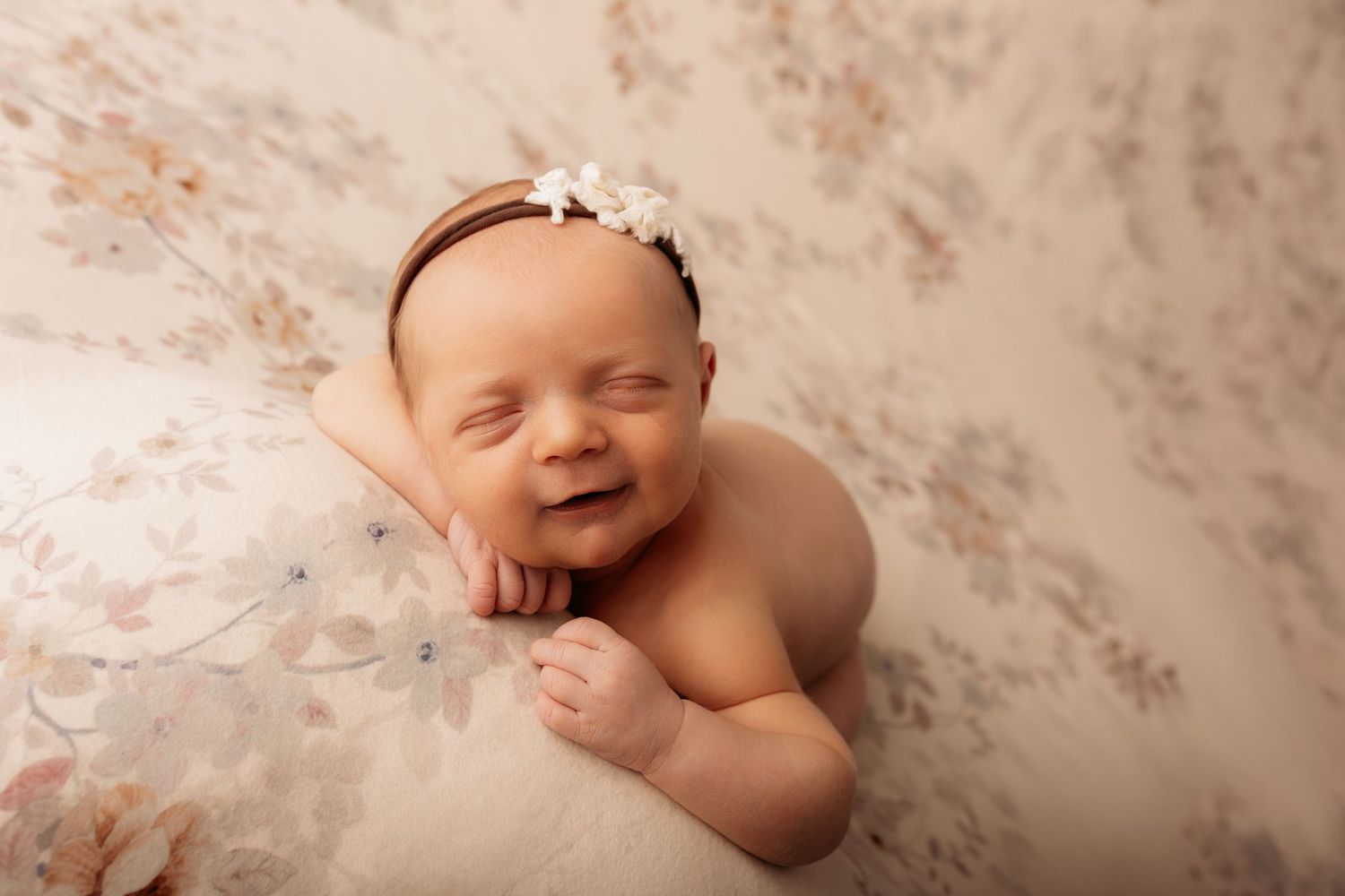 Smiling newborn baby girl wearing a white floral headband, posed on a soft cream floral blanket.