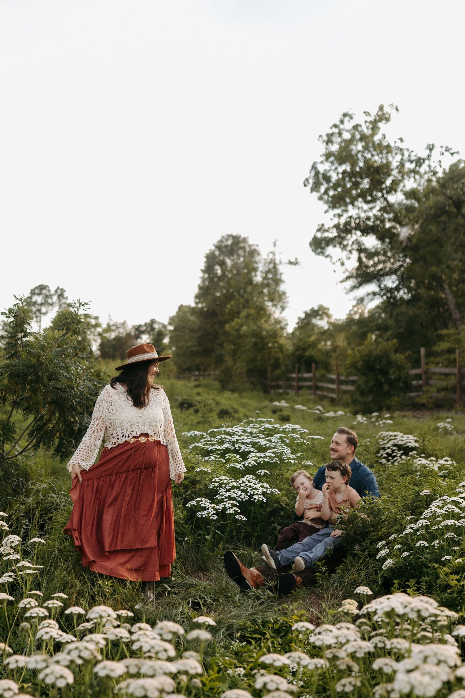 A bohemian family sits among wildflowers in a lush green field, with a woman in a red skirt and hat smiling at her partner and two babies.
