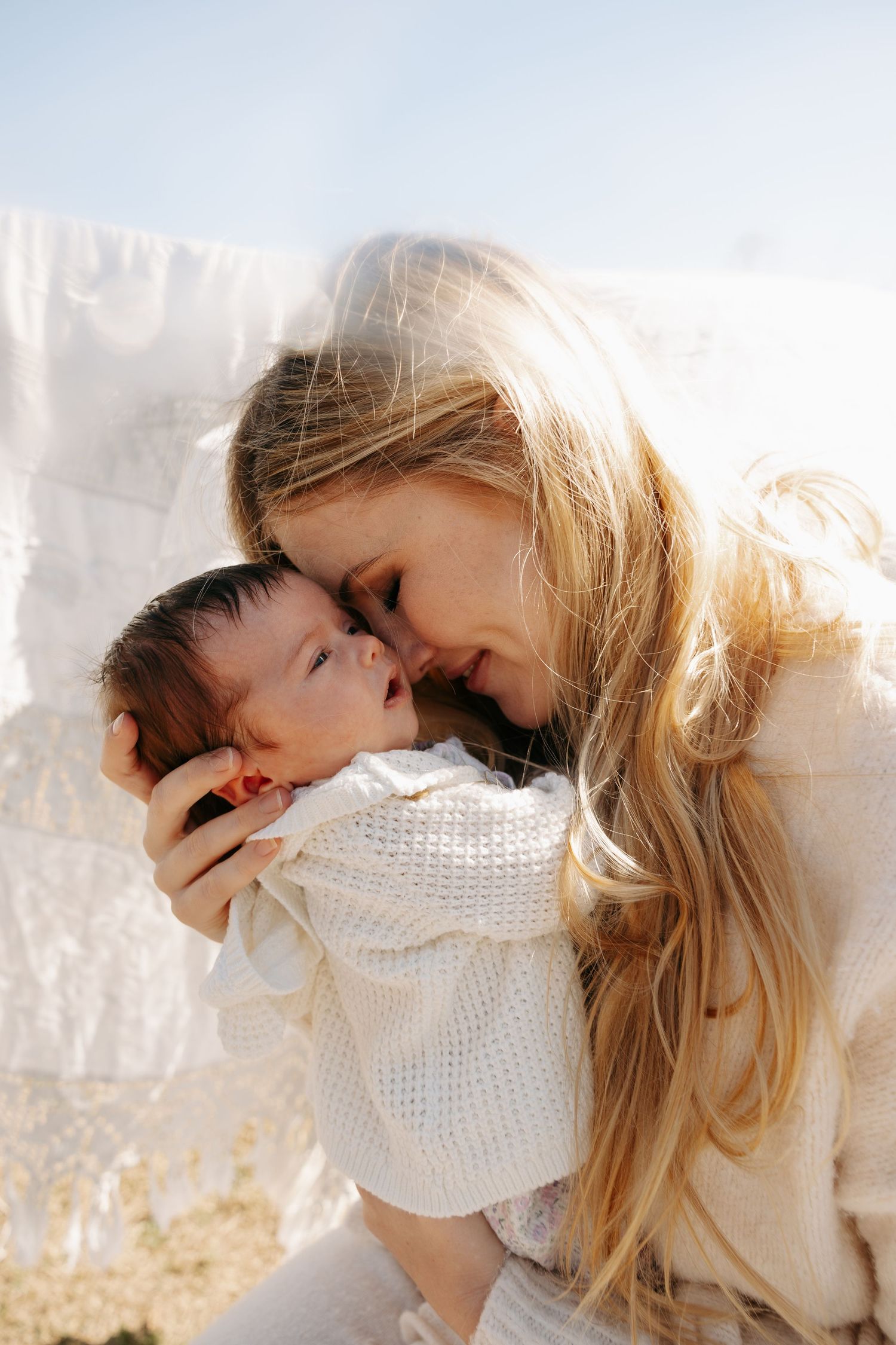 A smiling blonde mother tenderly holds her newborn baby face-to-face in soft natural sunlight outdoors.