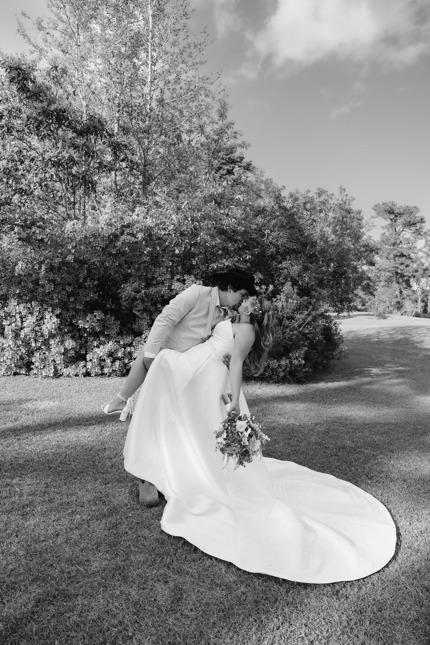 Black and white photo of groom dipping bride for a kiss outdoors with lush garden backdrop.