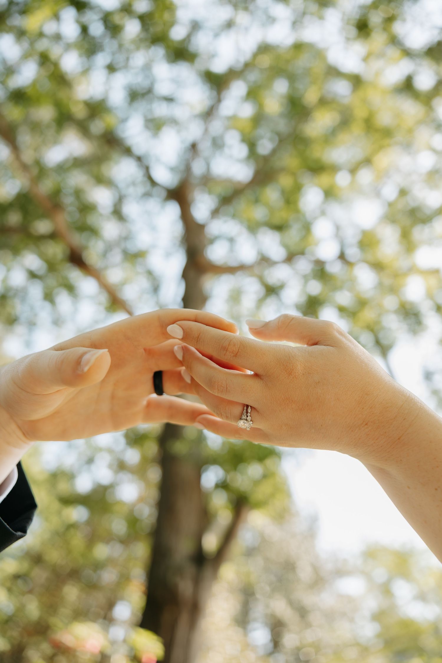 Two hands touching fingertips outdoors, one wearing a diamond engagement ring, with blurred green trees in background.