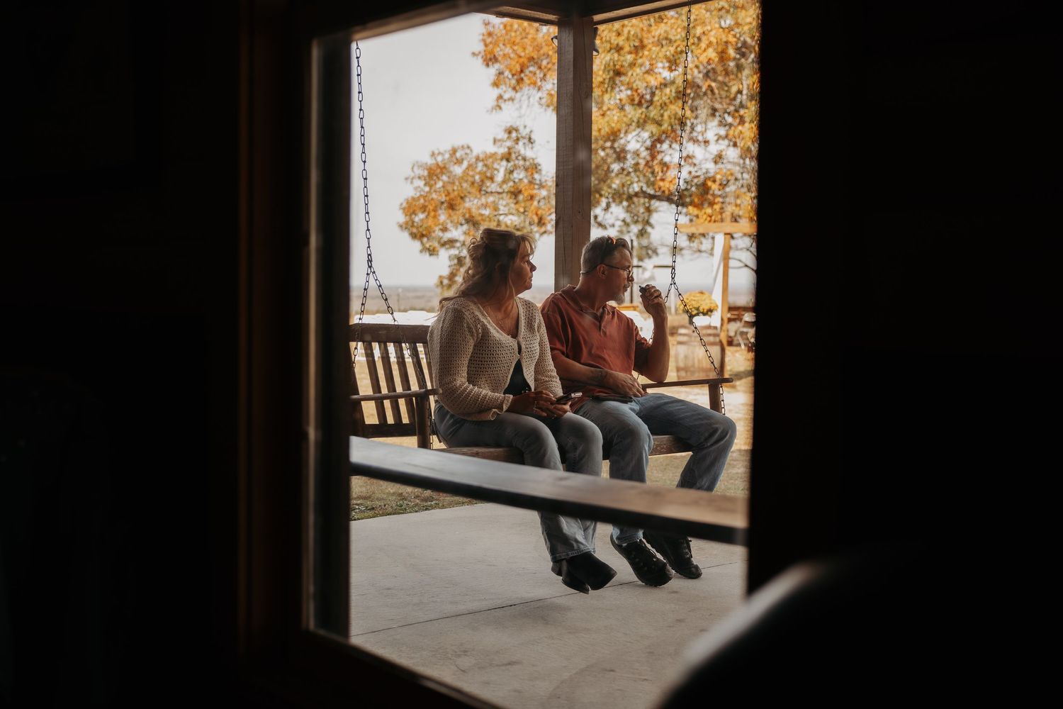 Two women sitting together on a porch swing, surrounded by autumn foliage, viewed through a window frame.