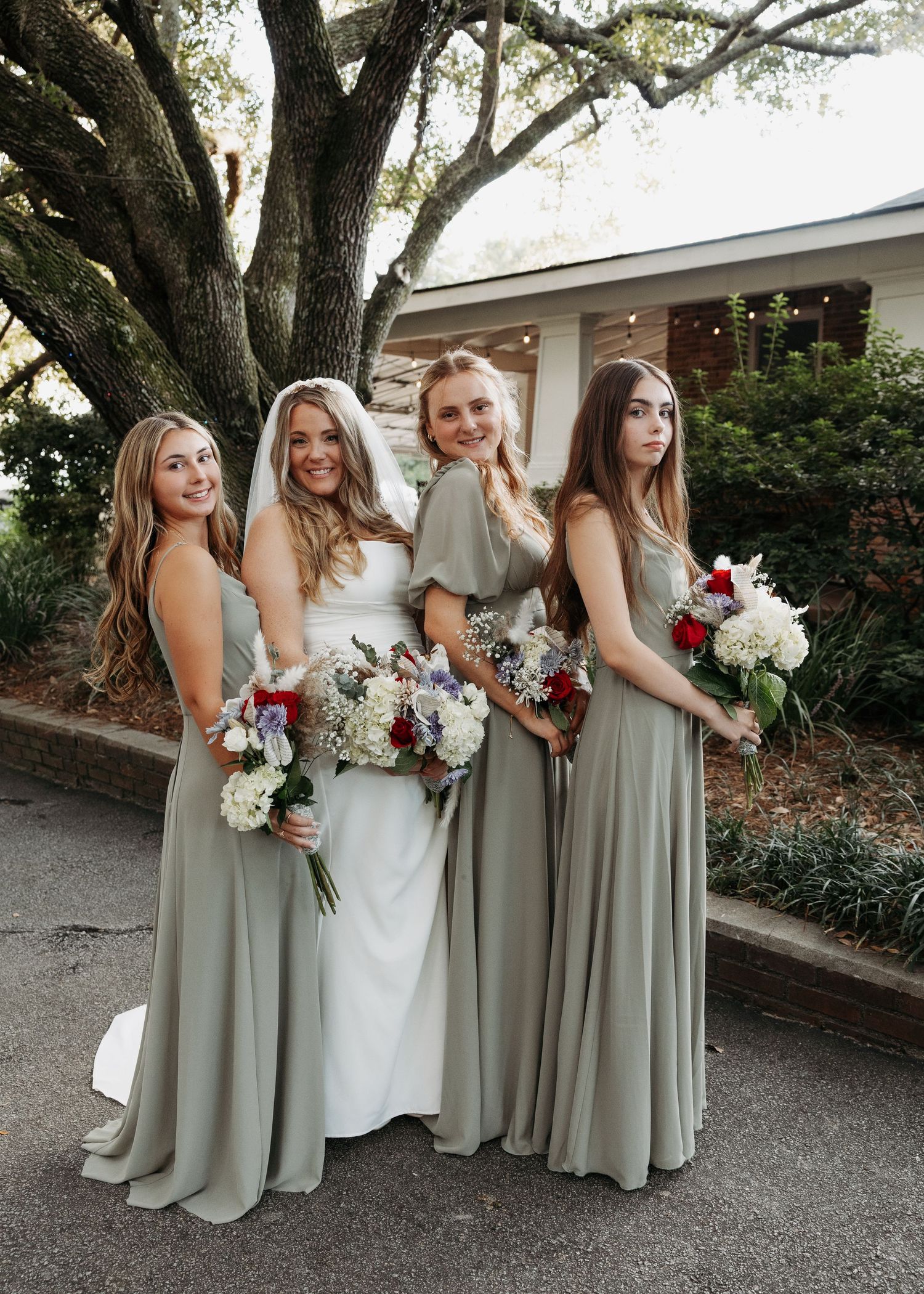 Bride with three bridesmaids in sage green gowns holding red and white floral bouquets.
