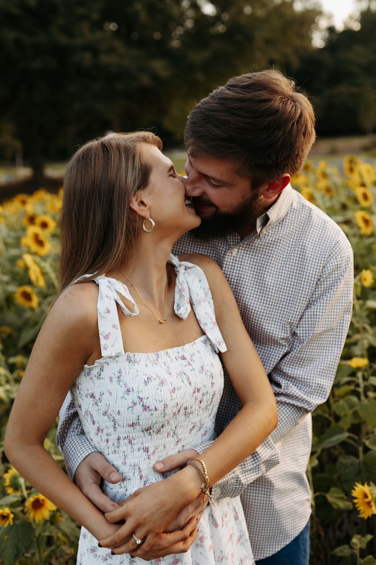 Couple sharing a romantic moment in a sunflower field, man embracing woman in floral dress at golden hour.