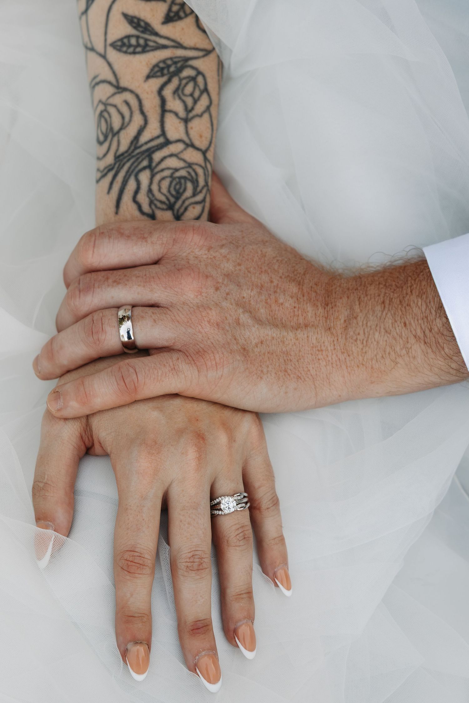 Close-up of newlywed couple's hands showing wedding rings, with a rose tattoo visible on the bride's arm.