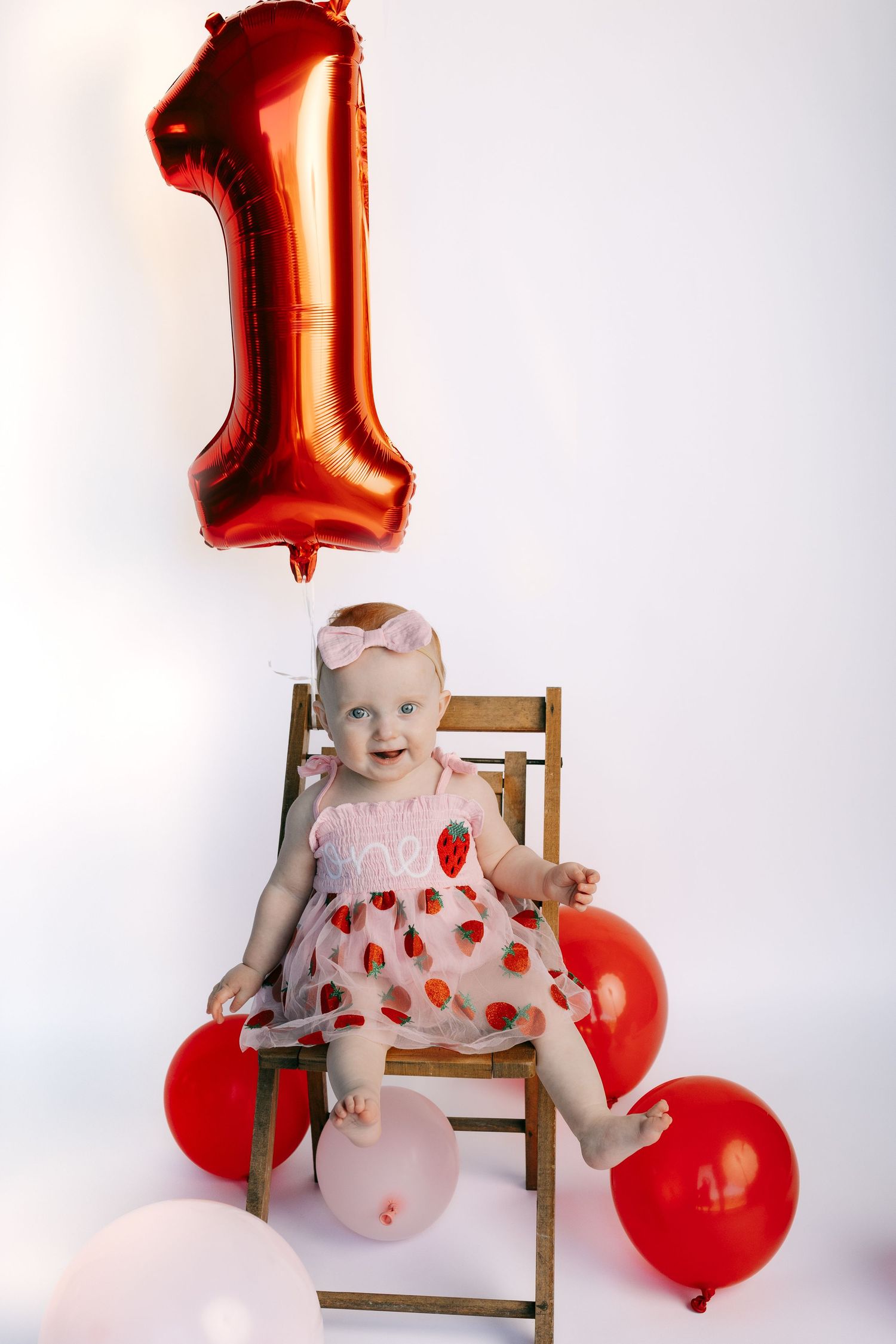 Baby girl in pink strawberry dress sits on chair with red number 1 balloon celebrating her first birthday.