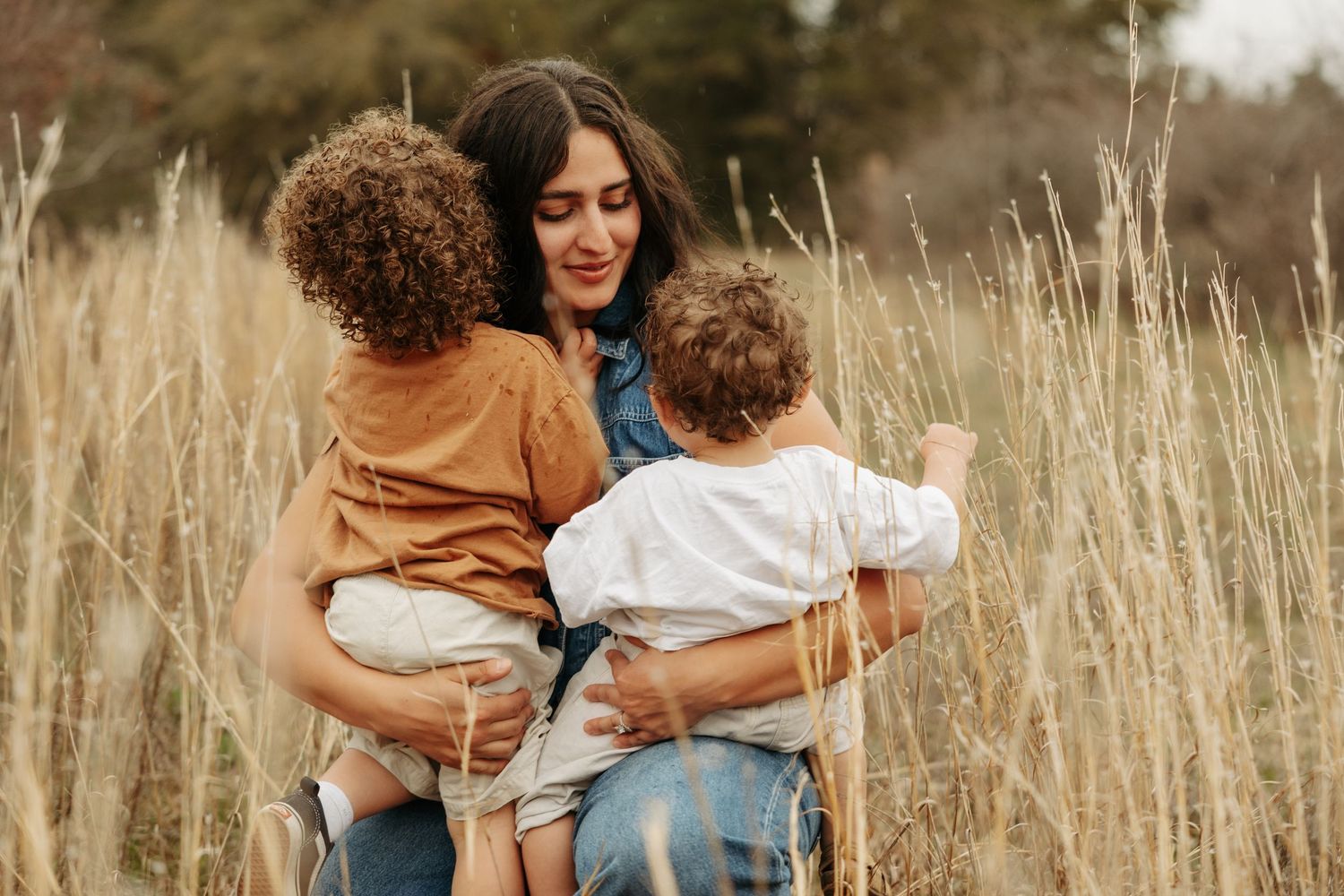 A mother holds two young curly-haired children in a golden field of tall grass during a warm outdoor family photo session.