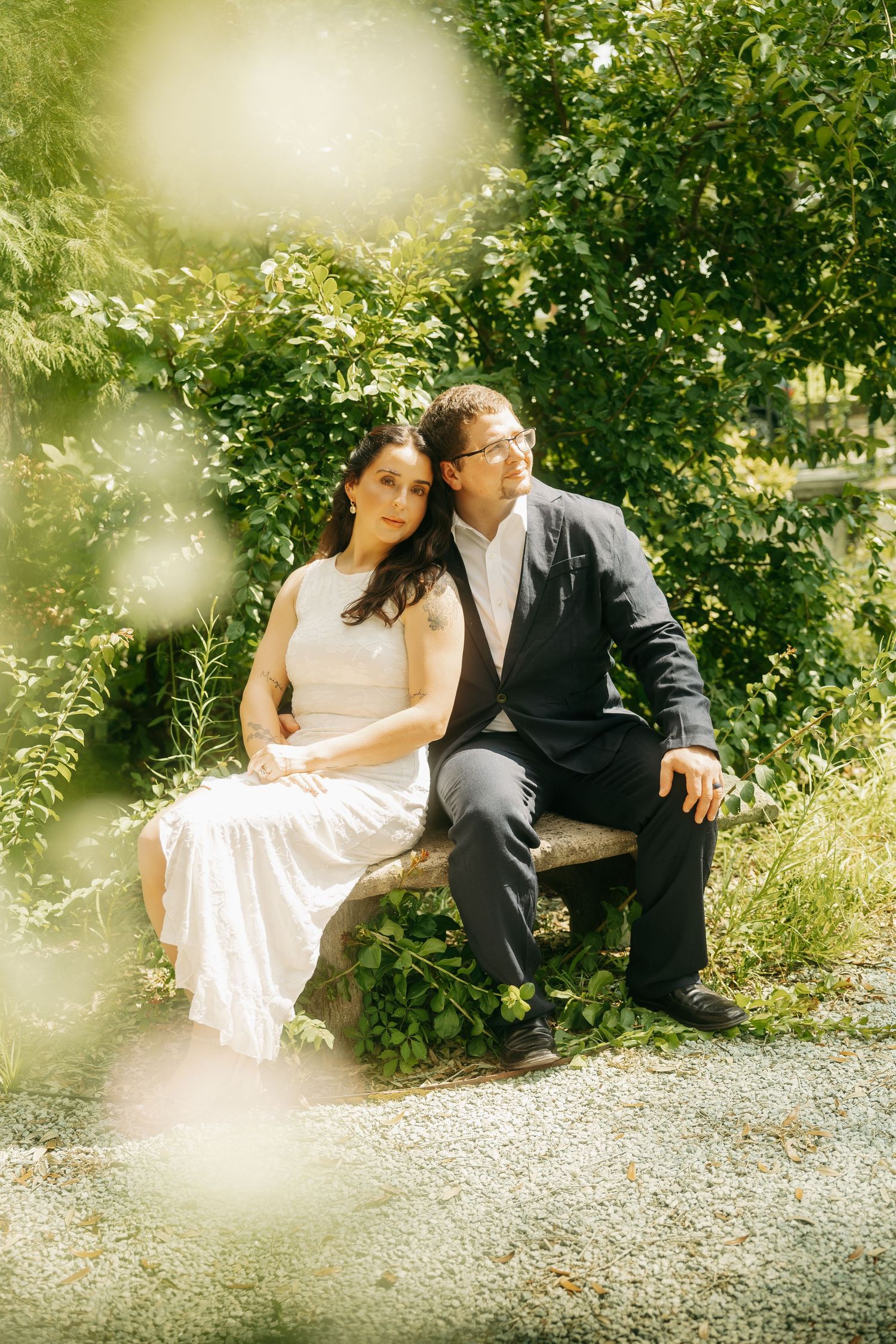 Couple in wedding attire sit closely together on a stone bench surrounded by green foliage in sunlight.
