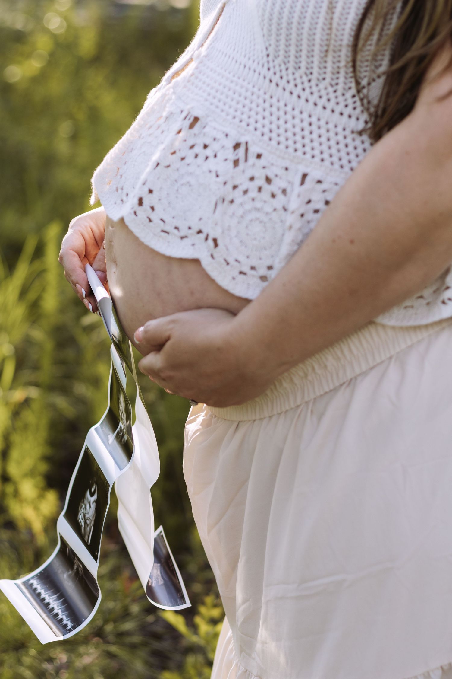 Pregnant woman in white lace dress holding ultrasound photos outdoors in sunlit grassy setting.