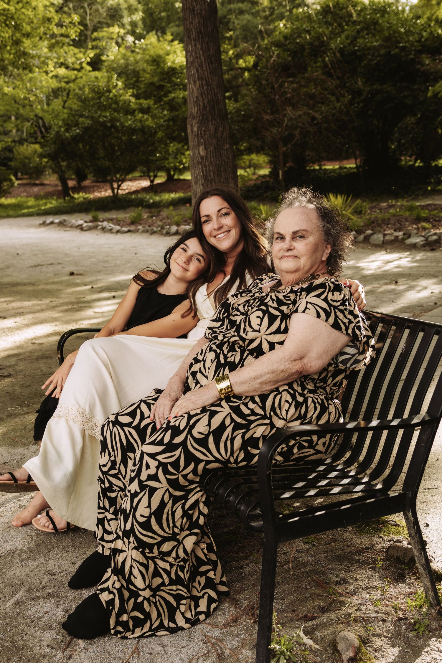 Two people pose together on a park bench under leafy trees during a sunny outdoor photo session.