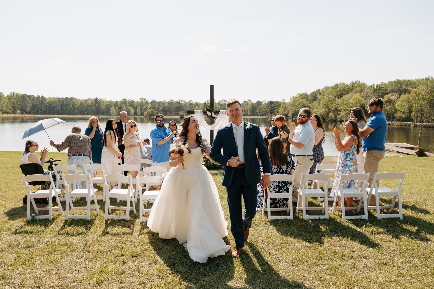 Happy newlyweds walk down outdoor aisle by lakeside as guests celebrate their wedding ceremony.