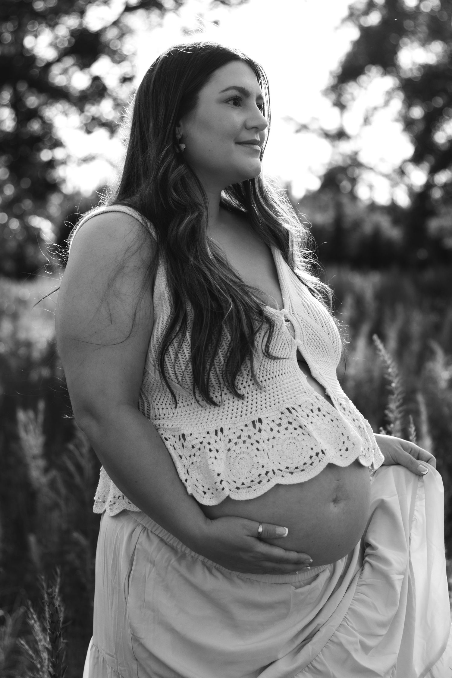 Black and white maternity photo of a woman in crochet top holding her baby bump outdoors.