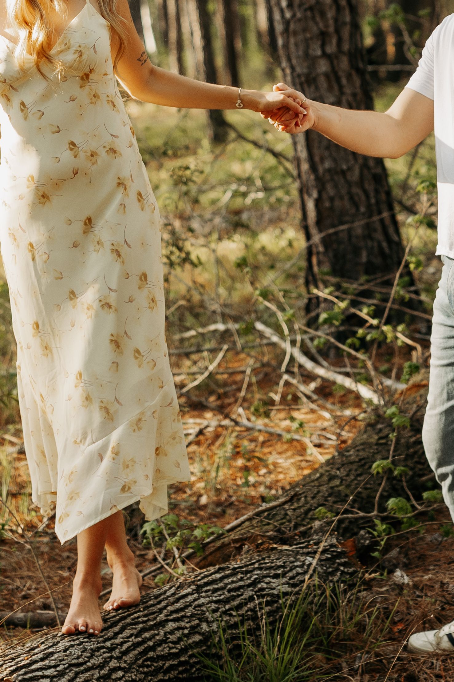 A woman in a floral dress balances barefoot on a log in the forest while holding hands with a companion.