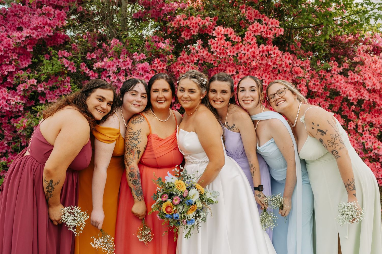Bride and six bridesmaids in rainbow-colored gowns smiling together in front of pink blooming flowers.