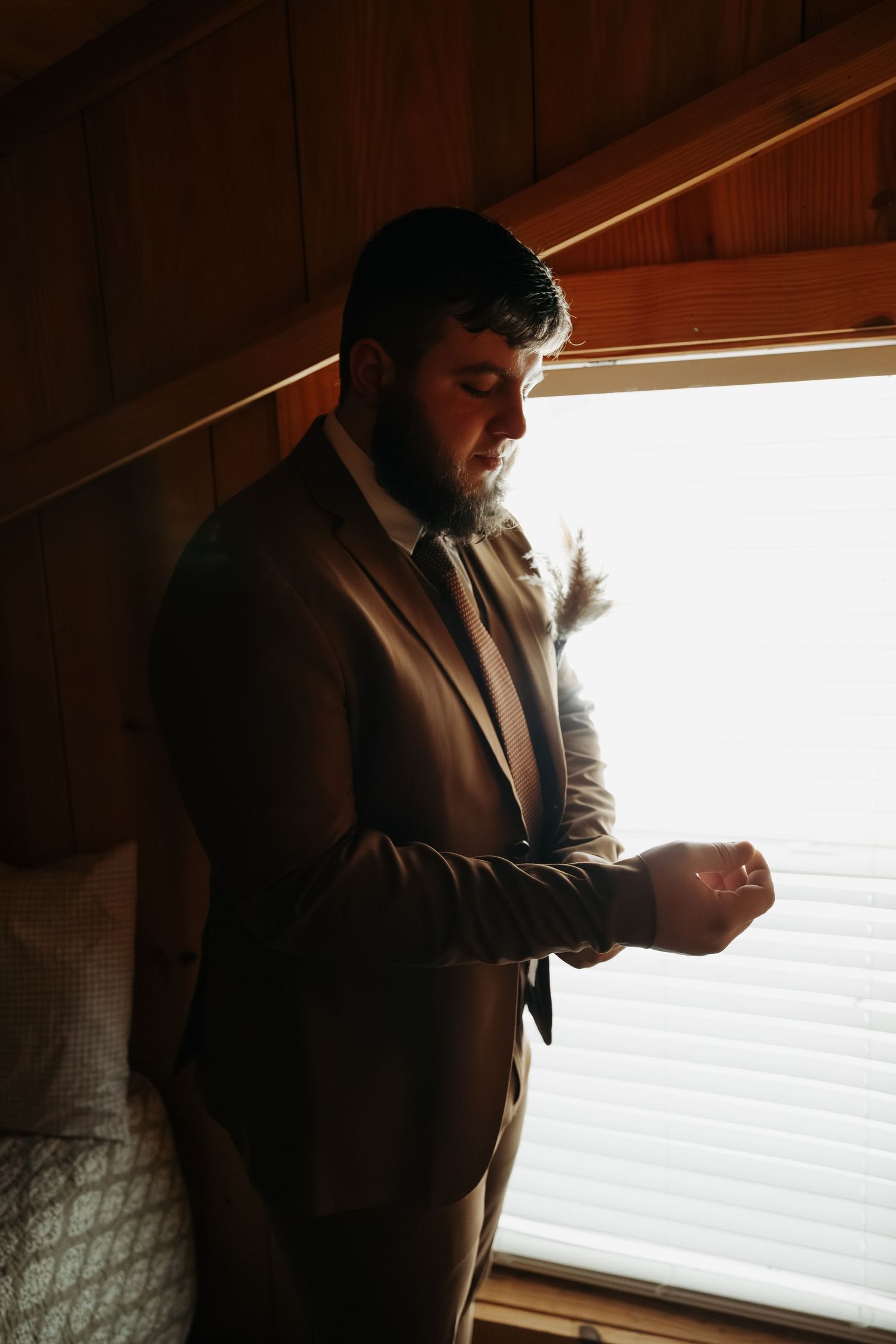 Groom in brown suit adjusting cufflinks by window on wedding day.