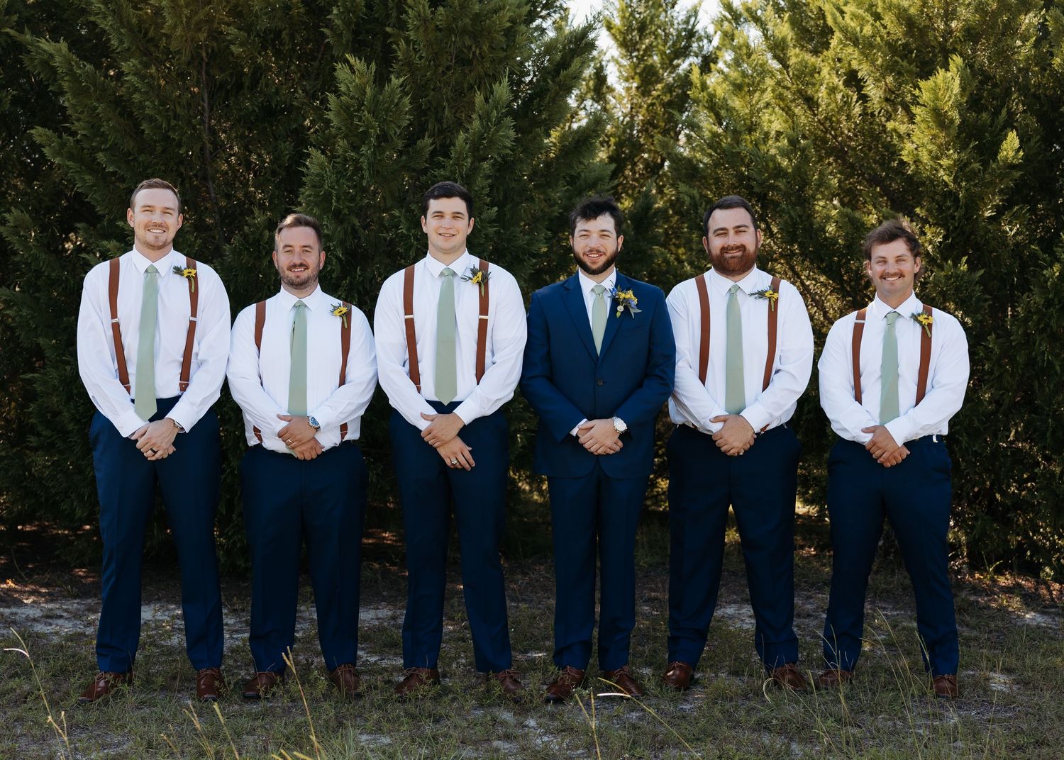 Six groomsmen in navy suits and brown suspenders pose outdoors before a wedding ceremony.