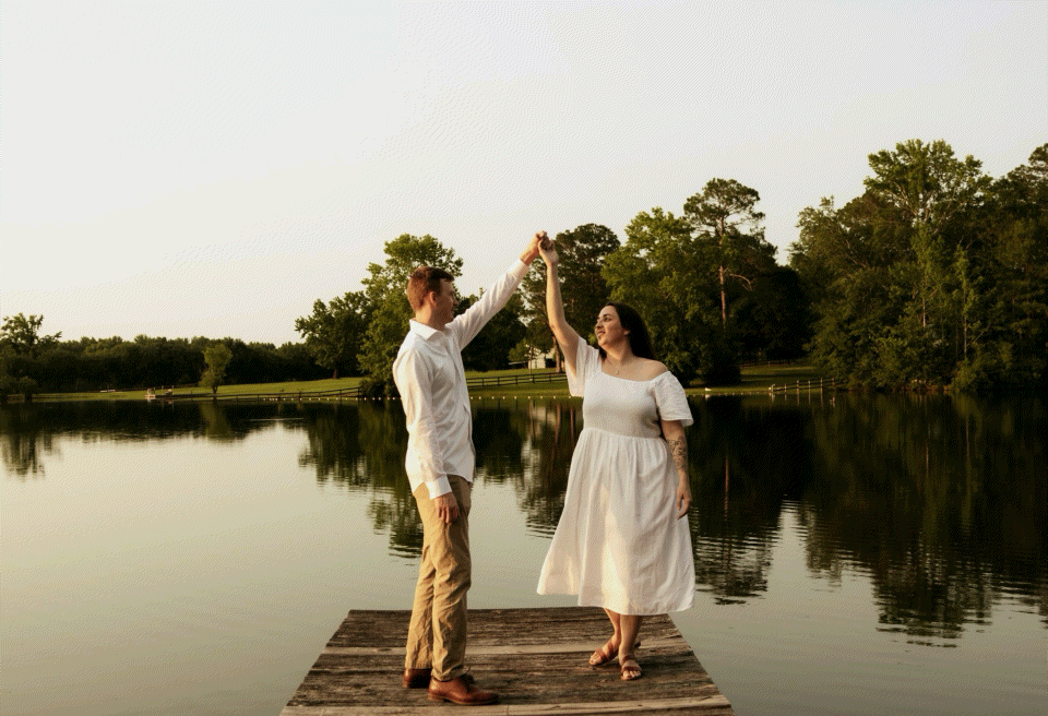 Couple dancing on a wooden dock by a serene lake at sunset, surrounded by lush green trees.