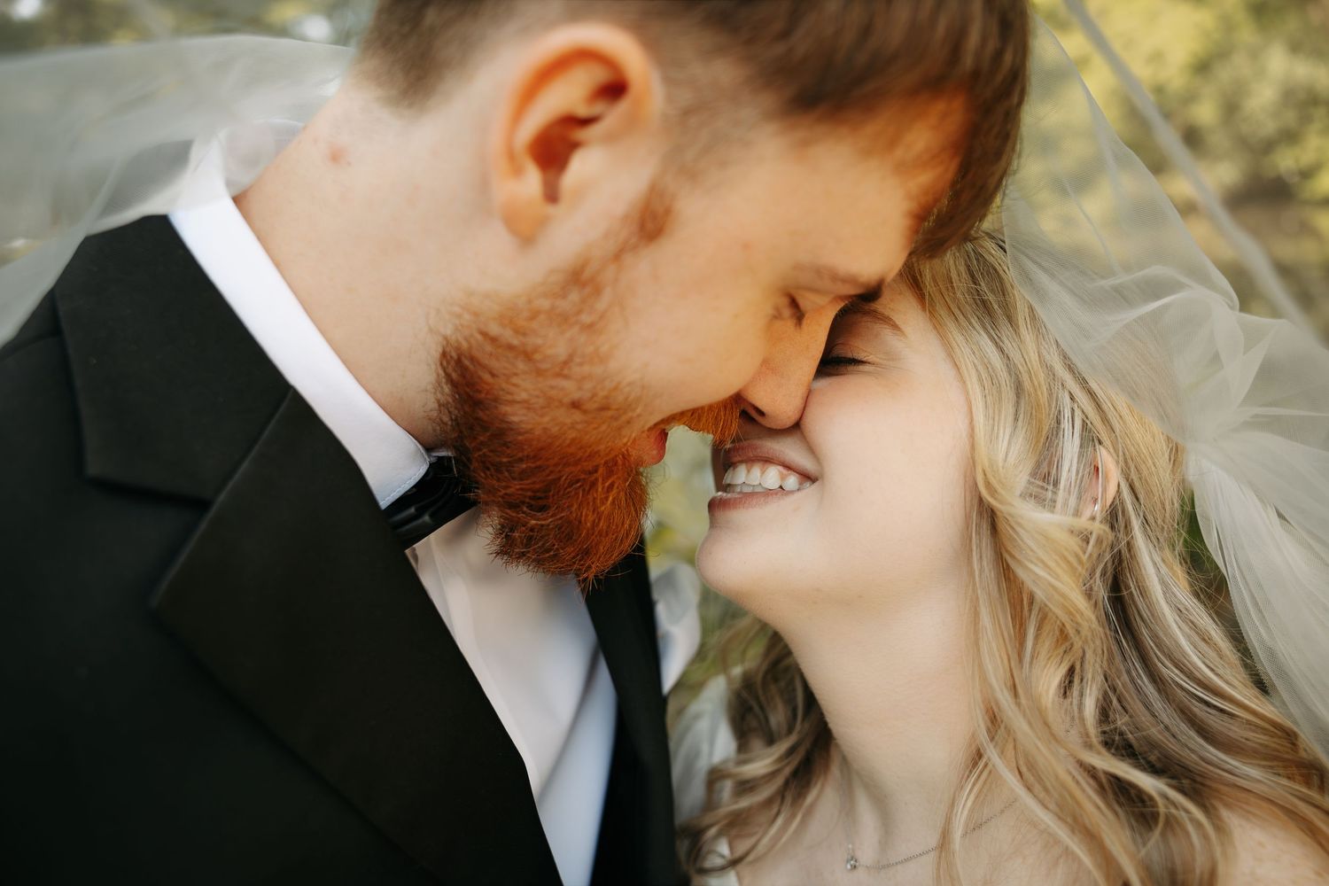A bride and groom share an intimate nose-to-nose moment, smiling tenderly under a wedding veil outdoors.