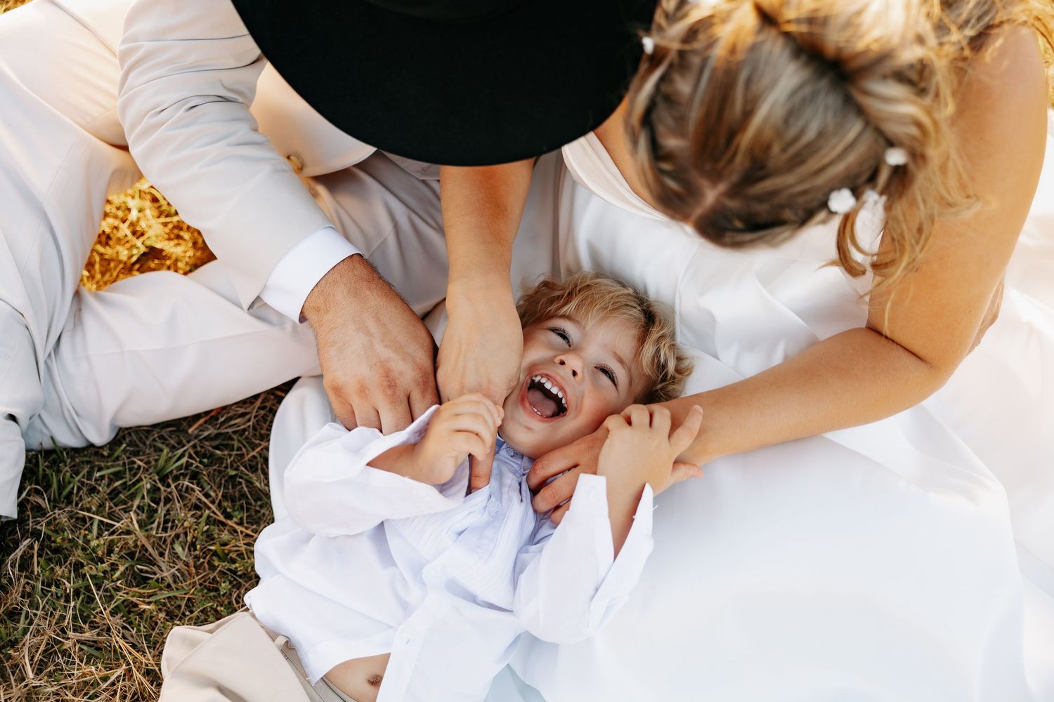 Bride and groom laughing while tickling a giggling child laying on the grass at a wedding.