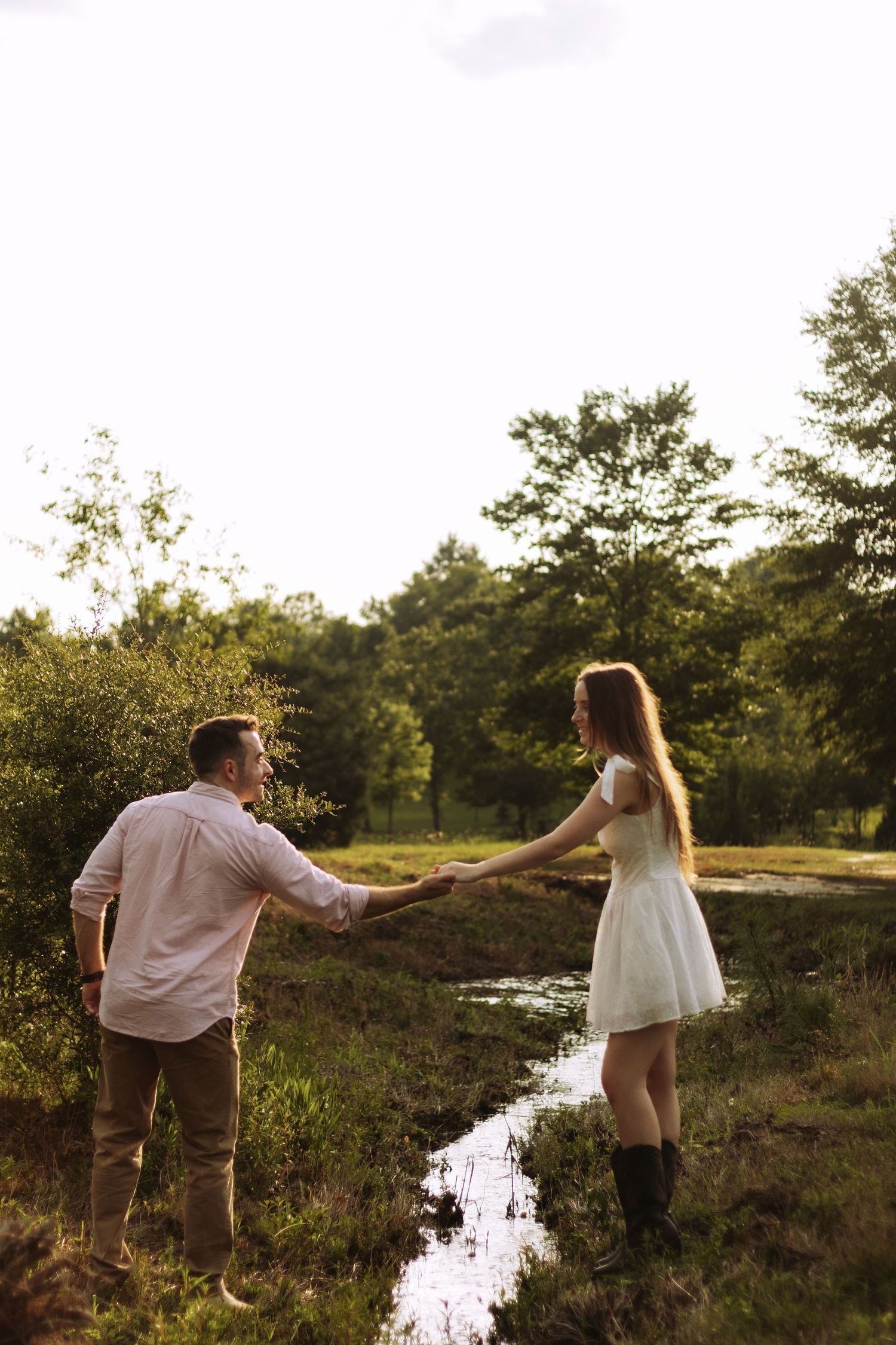 Couple holding hands by a stream in a sunlit meadow, woman in white dress and boots, man in casual shirt and khakis.
