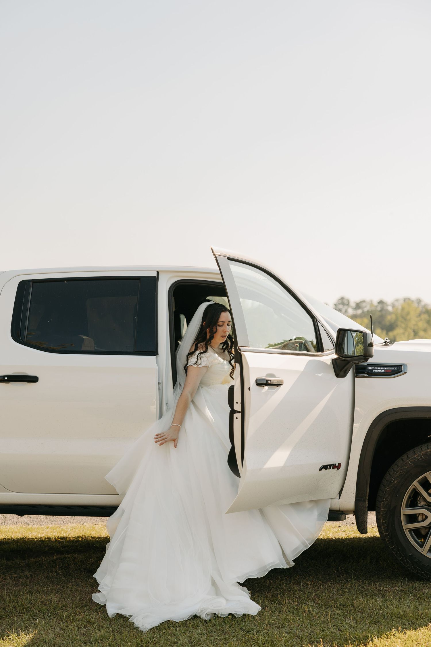 Elegant bride in flowing white gown steps out of white pickup truck on sunny wedding day.